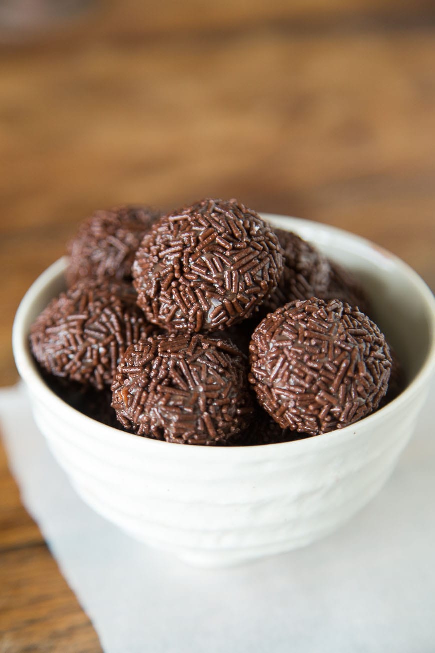 A white bowl of Brigadeiro, a Brazilian Chocolate Bonbon coated in chocolate sprkinles with the background blurred out.