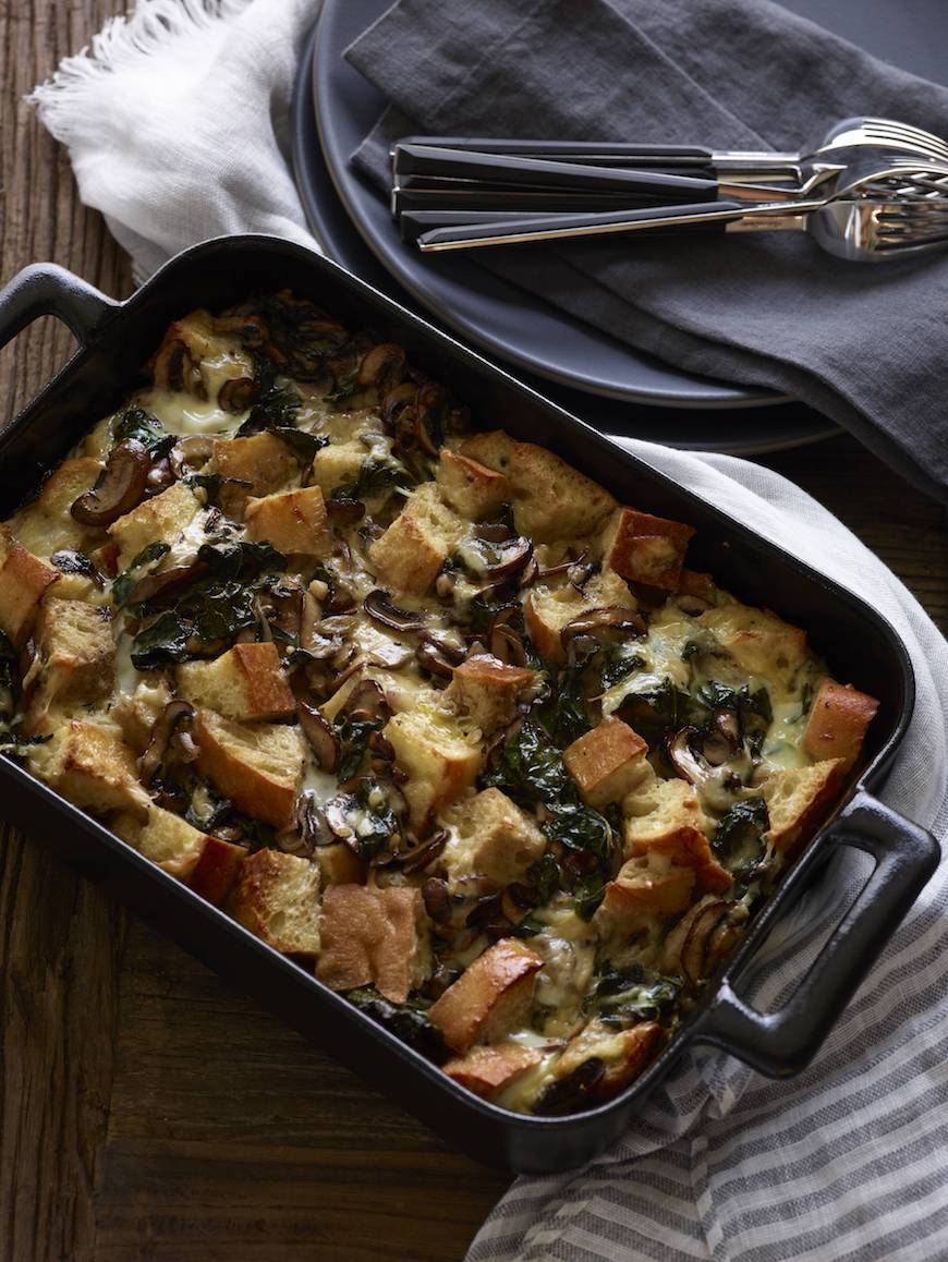 A black rectangular baking dish wrapped in a white kitchen towel with freshly baked Kale and Mushroom Bread Pudding with some black plates, black kitchen towels and forks placed next to it.