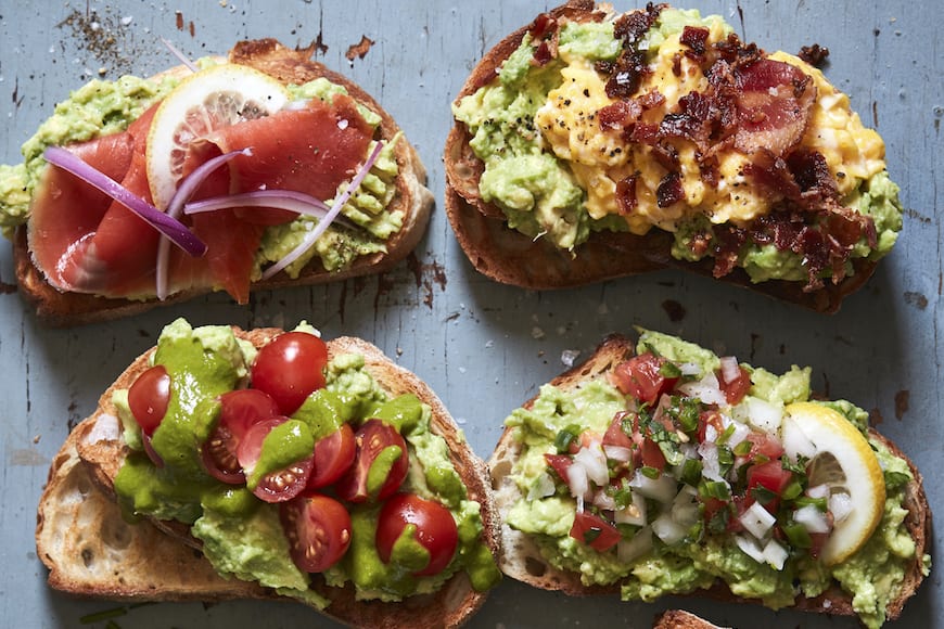 multiple slices of avocado toast with various toppings laid-out on a blue counter.