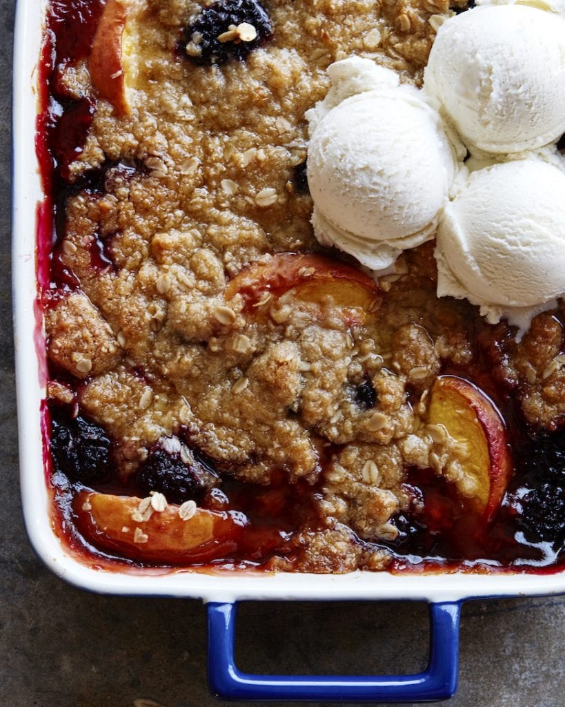 A tight shot of a bubbling peach and blackberry crisp in a cobalt blue rectangular ceramic baking dish with three scoops of vanilla ice cream in the top right corner.
