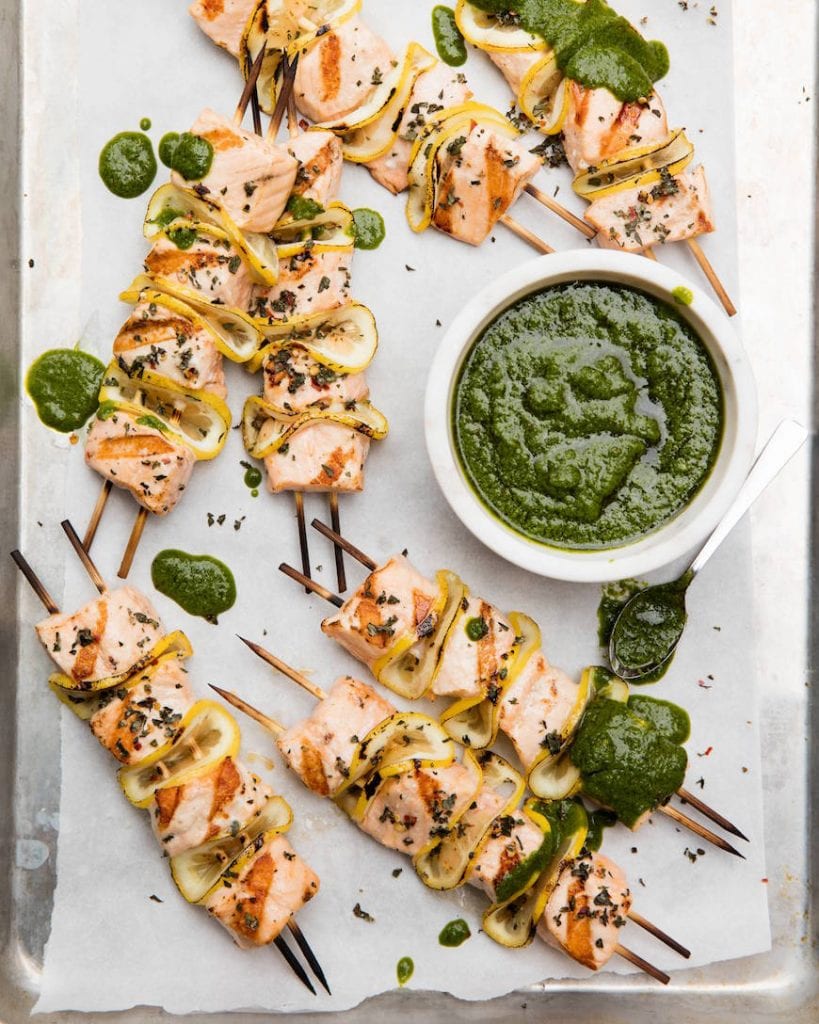 6 salmon skewers scattered on a baking sheet with parchment paper and drizzled with a basil vinaigrette. The skewers have grilled salmon chunks skewered between folded lemon slices. And a small bowl of basil vin for serving sits on the middle right of the pan.